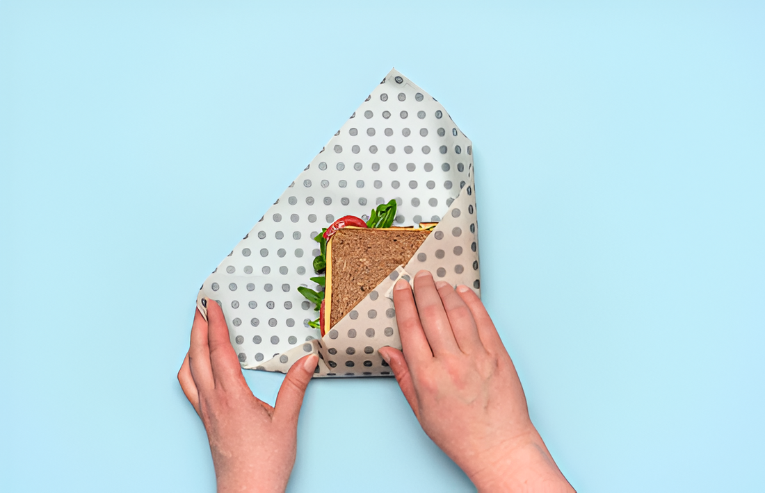 A pair of hands wrapping a sandwich with beeswax wrap on a light blue background